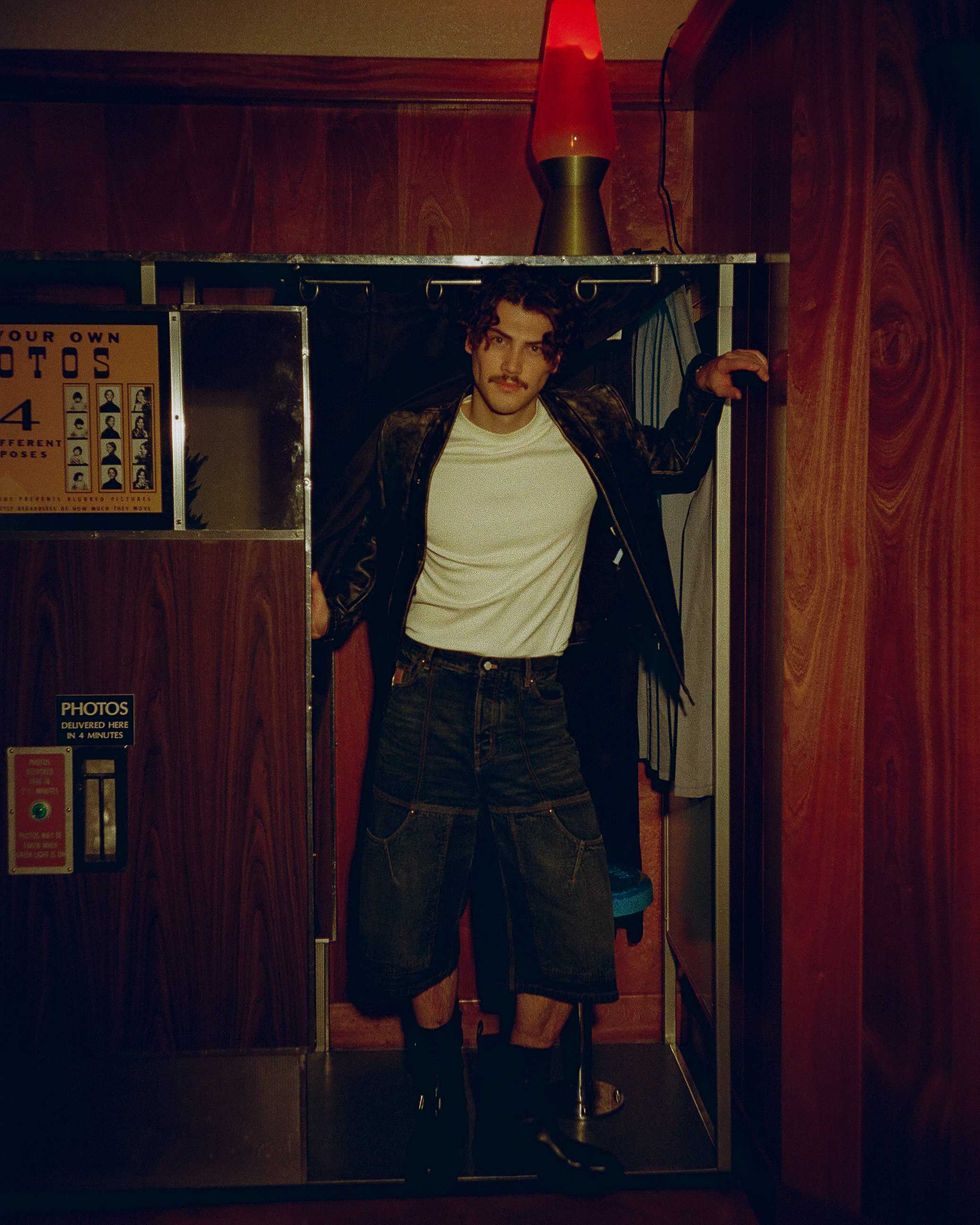 Jack Innanen in a white t-shirt, jean shorts, and leather jacket, standing in a Photo Booth.