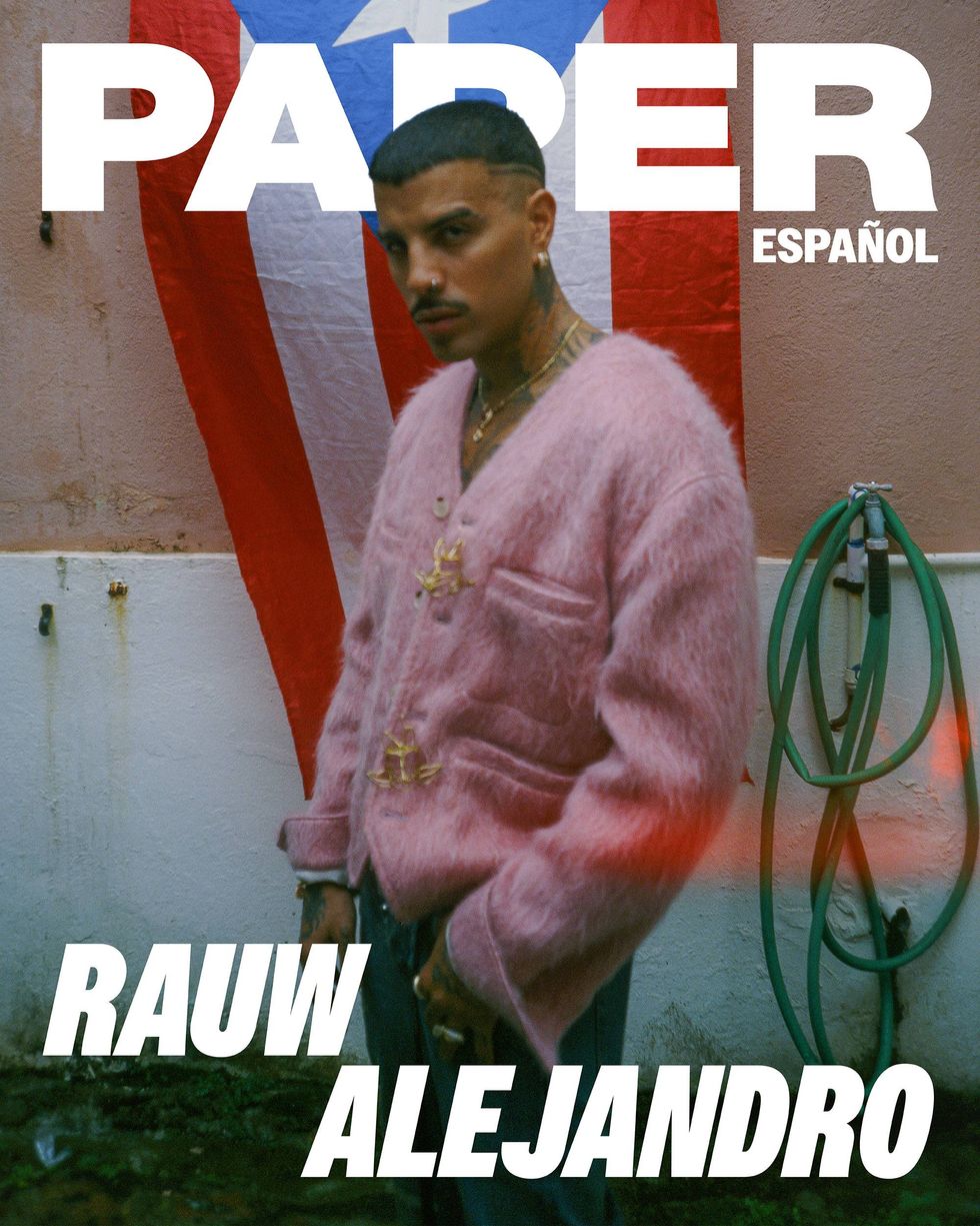Rauw Alejandro stands in front of a Puerto Rican flag in pink mohair cardigan and jeans.